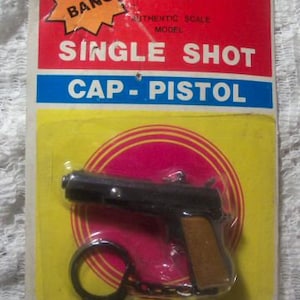 Op de afbeelding: Een zwart speelgoed-kapselpistool met een bruine handgreep en een zilveren sleutelhanger eraan. De verpakking is rood, geel en blauw met de tekst "Big Bang", "Single Shot" en "Cap-Pistol".