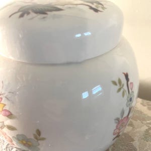May include: A white ceramic jar with a lid, decorated with floral patterns in pink, yellow, and blue. The jar has a rounded shape and a glossy finish, suitable for decorative or storage purposes.