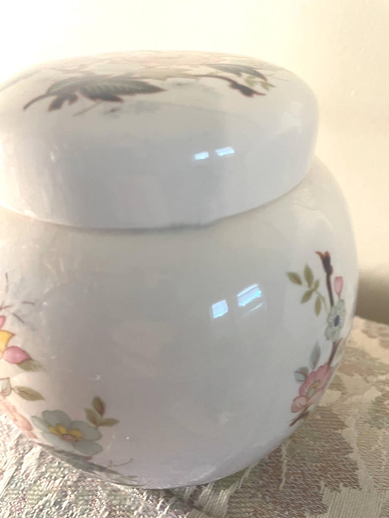 May include: A white ceramic jar with a lid, decorated with floral patterns in pink, yellow, and blue. The jar has a rounded shape and a glossy finish, suitable for decorative or storage purposes.