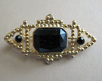 May include: A gold-toned brooch featuring a black faceted gemstone in the centre. The brooch has a geometric design with small black gemstones on either side.