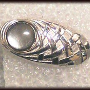May include: Silver-toned oval brooch with a central, circular, gray stone. The brooch features a woven design on one side and a smooth, polished finish on the other. The brooch is approximately 1.5 inches long.