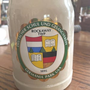 May include: A beige ceramic beer stein featuring a circular emblem with a scalloped gold border. The emblem displays the text DEUTSCHER SCHUL UND GESANGVEREIN, ROCKAWAY TWP., 1895, and GERMANIA PARK USA, surrounding a shield with a flag and book graphic.