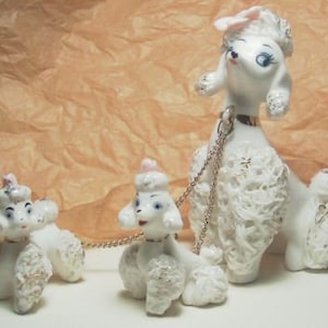 May include: Three white porcelain poodle figurines with pink bows. The largest poodle is standing and has a gold chain around its neck. The two smaller poodles are sitting and are connected to the larger poodle by the chain.