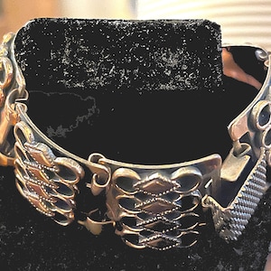 Può includere: Un braccialetto d'argento con un design dettagliato e intricato. Il braccialetto è realizzato con pezzi collegati e intrecciati che formano un motivo ripetitivo.