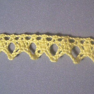May include: Close-up of a length of yellow crochet lace. The lace has a scalloped edge with a repeating pattern of openwork shapes. The background is a solid, muted gray color.