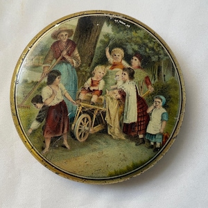 Op de afbeelding: Ronde, antieke blikken doos met een geschilderde scène van spelende kinderen in de buitenlucht. De afbeelding toont een vrouw met een hark, kinderen met een kruiwagen en andere kinderen in een bosrijke omgeving. De doos heeft een goudkleurige rand.