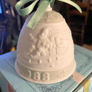 May include: A white ceramic bell with a pale green ribbon. The bell has a relief design of a person and buildings, with the year "1988" at the base. The bell is on a light blue box.