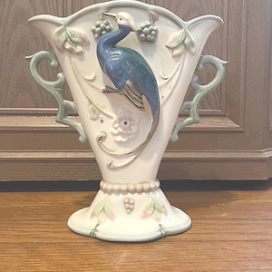 May include: A cream-colored ceramic vase with a blue and green bird design. The vase has a flared top with decorative handles and leaf accents. The base features a floral and grape design.
