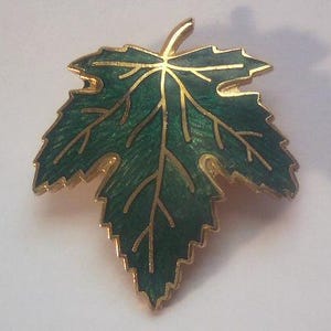 May include: A gold-toned leaf-shaped brooch with a detailed design. The brooch features a vibrant green enamel filling, with gold-colored veins and edges. The leaf has a textured appearance, adding depth to the design.