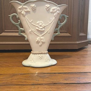 May include: A cream-colored ceramic vase with a flared top and base, featuring embossed grape and leaf designs. The vase has two light green handles and accents. The vase is approximately 10 inches tall.