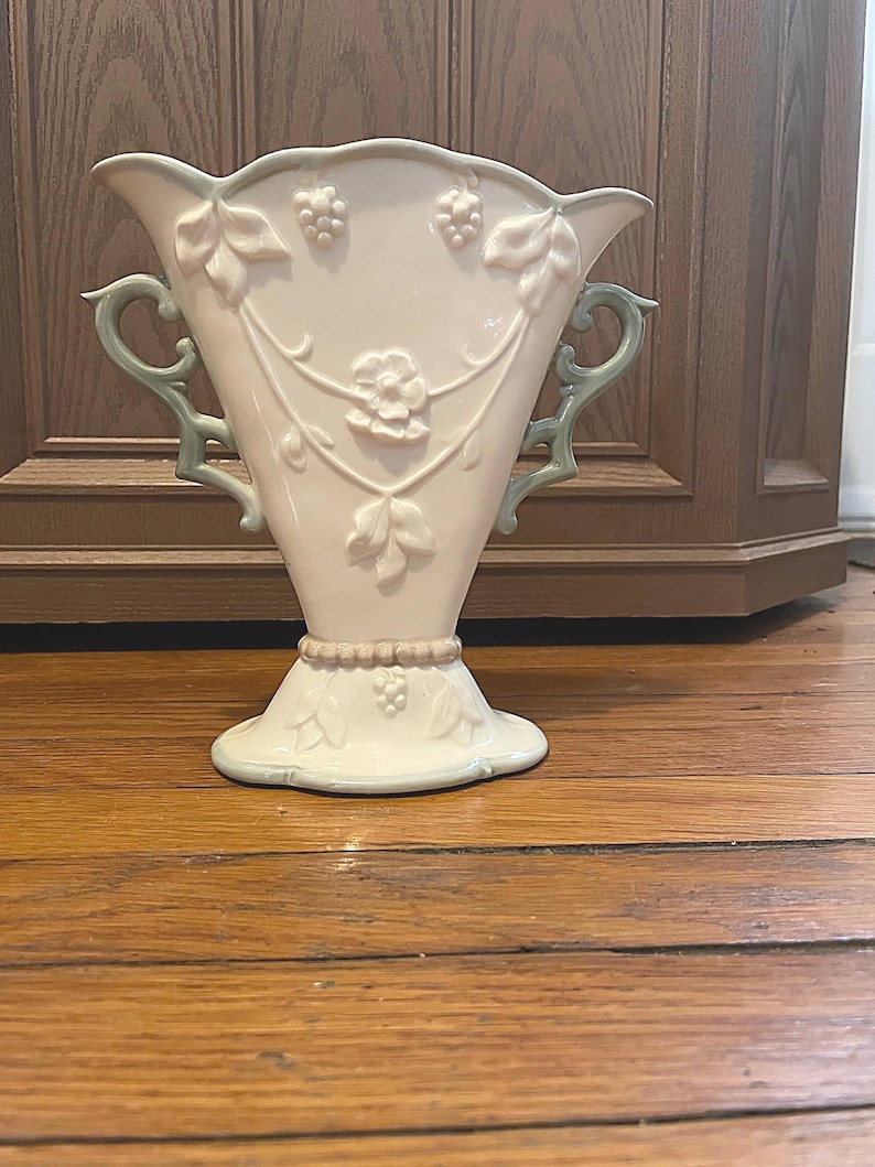 May include: A cream-colored ceramic vase with a flared top and base, featuring embossed grape and leaf designs. The vase has two light green handles and accents. The vase is approximately 10 inches tall.