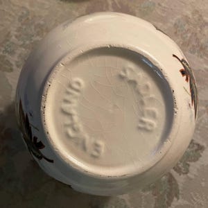 May include: White ceramic bowl with a floral design in shades of brown, green, and blue. The base of the bowl has the text "Ohio" and "213" embossed. The bowl has a cracked glaze.