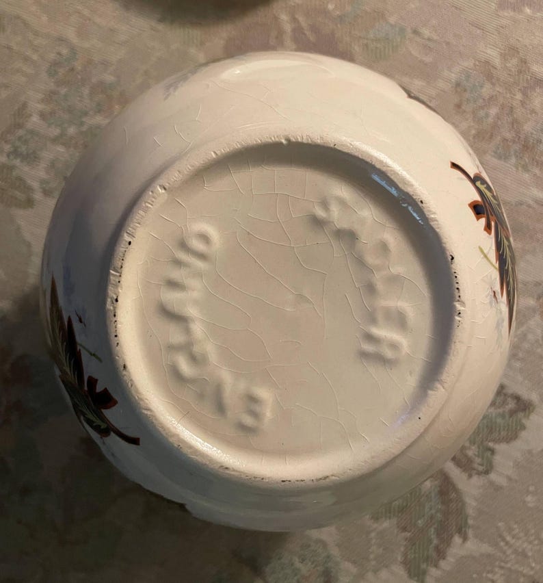May include: White ceramic bowl with a floral design in shades of brown, green, and blue. The base of the bowl has the text "Ohio" and "213" embossed. The bowl has a cracked glaze.