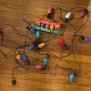 Pode incluir: Uma série de luzes de Natal vintage C-9½ com várias lâmpadas coloridas, incluindo vermelho, rosa, azul e amarelo. As luzes estão em um fio marrom e estão espalhadas em uma superfície de madeira. Uma caixa de lâmpadas de reposição também é visível.