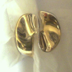 May include: A gold-colored ring with a unique, abstract design. The ring is made of two pieces that are joined together in the middle, creating a flowing, organic shape.