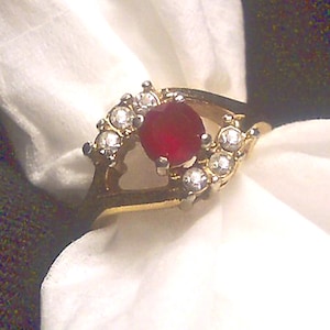 May include: A gold ring with a large red gemstone surrounded by smaller clear gemstones.