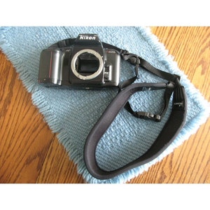 May include: Black Nikon N6006 35mm film camera with a black neck strap. The camera is on a light blue textured surface. The camera has a silver lens mount and the Nikon logo.