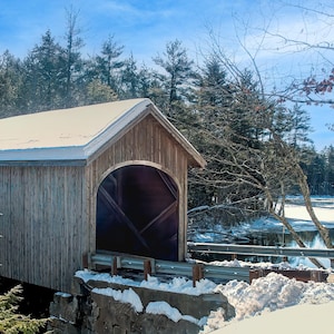 May include: A wooden covered bridge with a snow-covered roof, set against a backdrop of trees and a frozen lake. The bridge is made of wood and has a traditional design.