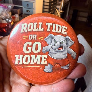 May include: A round, red button with the text "ROLL TIDE OR GO HOME" in white and yellow lettering. An angry cartoon elephant wearing red shorts is featured on the button. The button is being held.