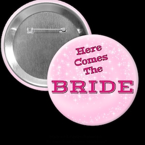Puede incluir: Un botón rosa claro con el texto "Here Comes The BRIDE" en negrita, rosa. El botón tiene un alfiler plateado en la parte posterior y un fondo brillante. Un segundo botón es parcialmente visible.