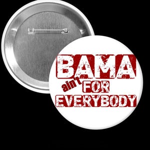 May include: A white button with the text "BAMA ain't FOR EVERYBODY" in distressed red lettering. The button has a metal pin back and a silver metal rim.