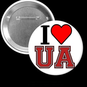 Puede incluir: Un botón blanco con el texto "I ♥ UA" en negro y rojo. El corazón es rojo, y las letras "UA" están en una fuente roja de estilo universitario con un contorno negro. Un segundo botón es visible en el fondo.