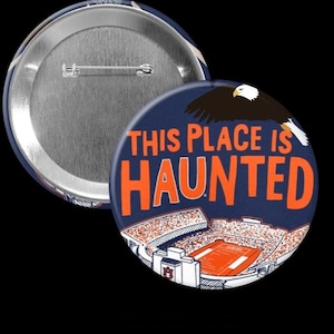 Puede incluir: Un botón azul marino con el texto "THIS PLACE IS HAUNTED" en naranja. El botón presenta una ilustración de un estadio y un águila volando. La parte posterior del botón es plateada.