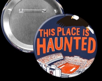 May include: A navy blue button with the text "THIS PLACE IS HAUNTED" in orange. The button features an illustration of a stadium and a soaring eagle. The back of the button is silver.