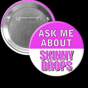 May include: A round button with a pink and white design. The top half is pink with the words "ASK ME ABOUT" in white. The bottom half is white with the words "SKINNY DROPS" in pink. The back of the button is silver.