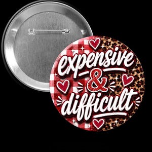 May include: A round pin-back button featuring a red and white checkered pattern on the left and a brown leopard print on the right. The text "expensive & difficult" is written in white script with a red glittery ampersand and small red hearts.