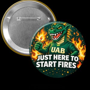 May include: A round button with a green dragon mascot holding a lit match, surrounded by flames. The text reads "UAB JUST HERE TO START FIRES". The button has a metallic pin back and a gold-colored rim.