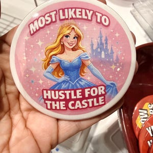 May include: Circular button with a princess illustration in a blue dress and tiara. The text "MOST LIKELY TO" and "HUSTLE FOR THE CASTLE" are in pink. A castle is in the background. The button has a white edge.
