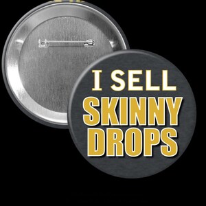May include: A round, black button with the text "I SELL SKINNY DROPS" in gold letters with a white outline. The button is set against a black background, with a silver pin back visible.