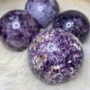 May include: Four purple and white Lepidolite spheres, each with a unique pattern of swirling colors and textures. The spheres are arranged on a white fluffy surface.