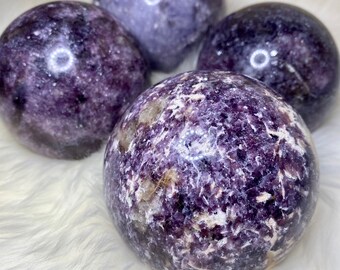 May include: Four purple and white Lepidolite spheres, each with a unique pattern of swirling colors and textures. The spheres are arranged on a white fluffy surface.