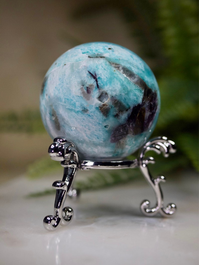 May include: A blue and white sphere with black and brown inclusions, resting on a silver metal stand with ornate details.