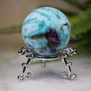 May include: A blue and green aquamarine sphere with purple inclusions, displayed on a silver stand.