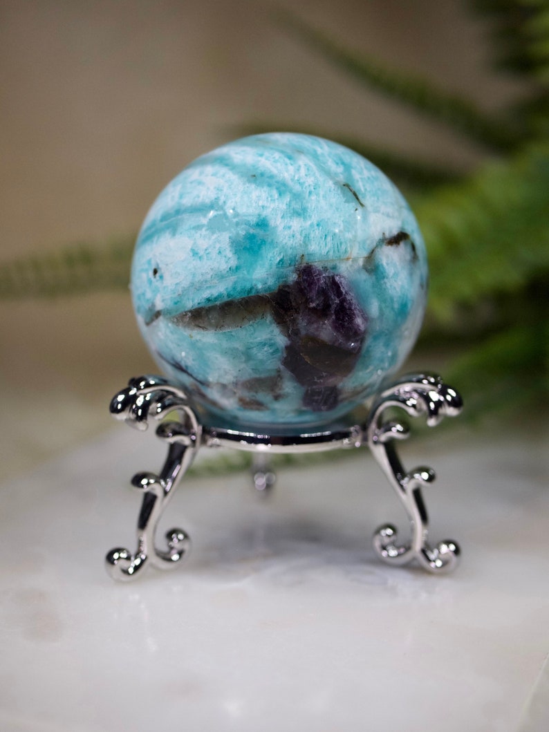 May include: A blue and green aquamarine sphere with purple inclusions, displayed on a silver stand.