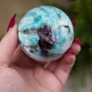 May include: A blue and green amazonite sphere with a purple amethyst inclusion. The sphere is smooth and polished.