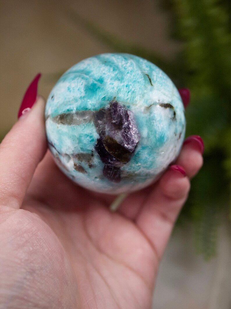 May include: A blue and green amazonite sphere with a purple amethyst inclusion. The sphere is smooth and polished.