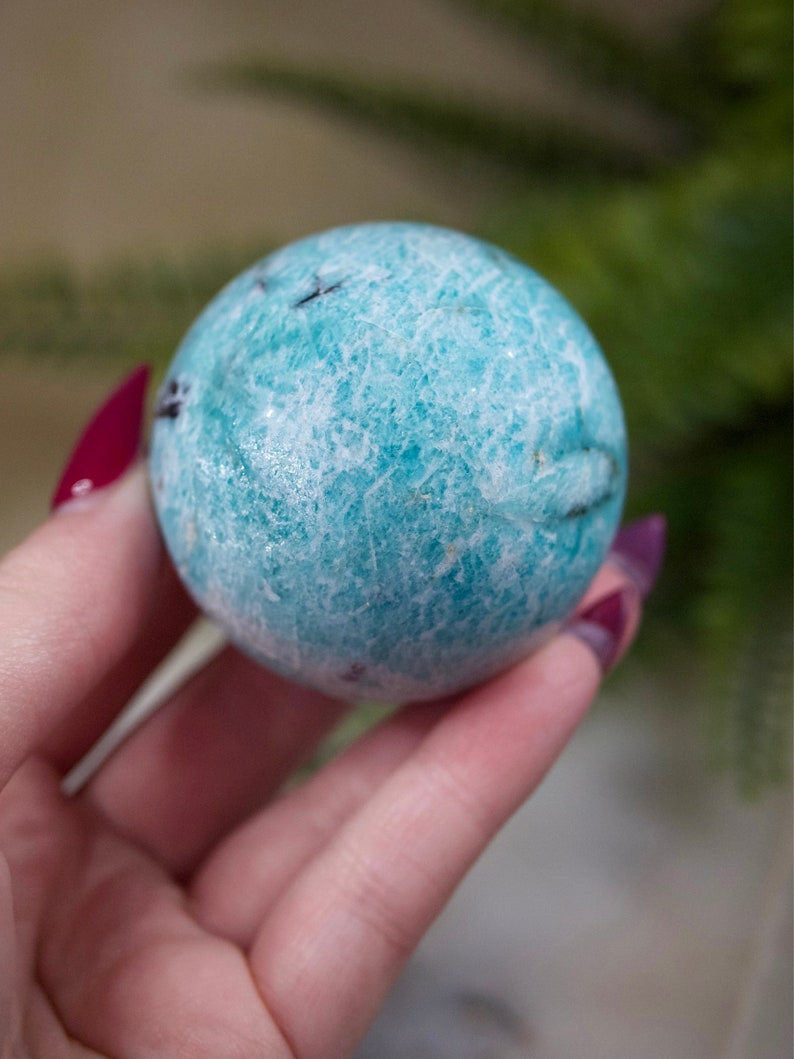 May include: A smooth, light blue and white sphere of Amazonite crystal. The sphere has a natural, textured surface.