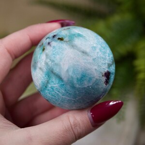 May include: A blue and white sphere with purple and green inclusions. The sphere is smooth and polished. It is being held in a hand.