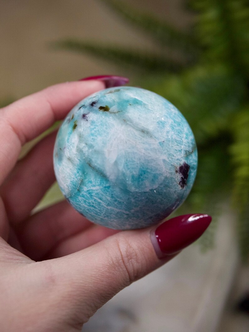 May include: A blue and white sphere with purple and green inclusions. The sphere is smooth and polished. It is being held in a hand.