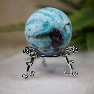 May include: A blue and green sphere with white and purple swirls sits on a silver stand with ornate legs. The sphere is a polished gemstone.