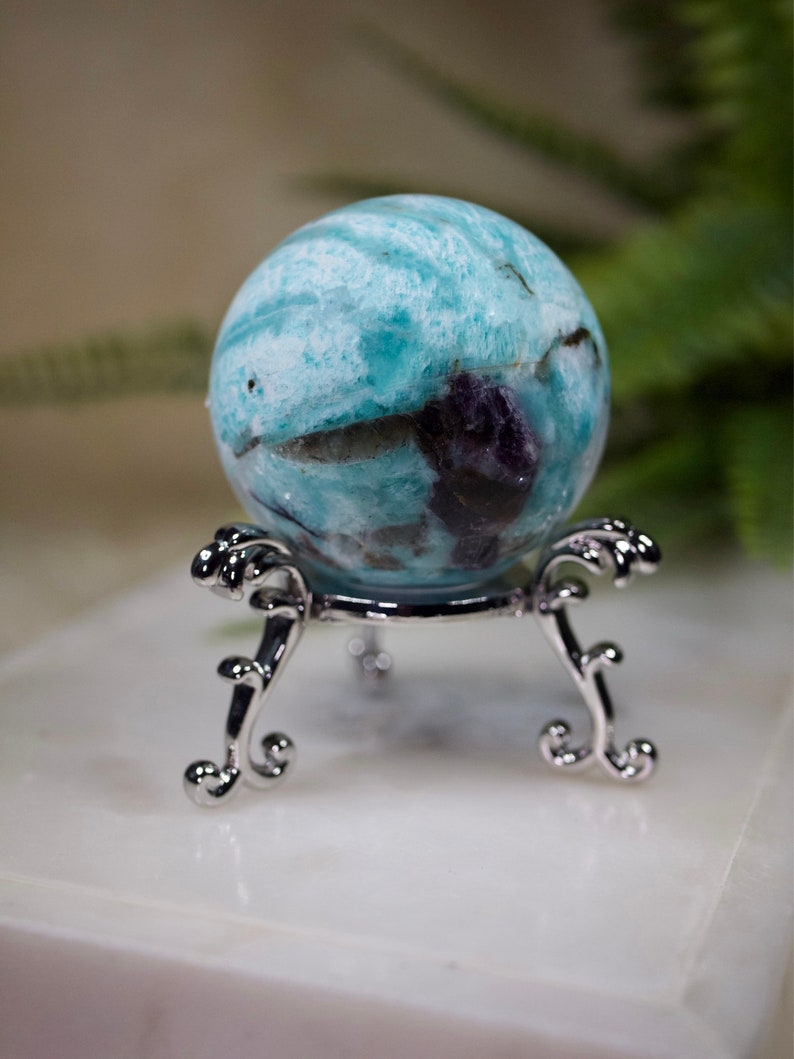 May include: A blue and green sphere with white and purple swirls sits on a silver stand with ornate legs. The sphere is a polished gemstone.