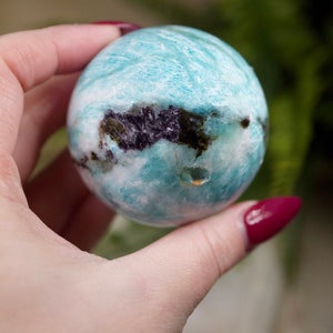 May include: A smooth, light blue and white Amazonite sphere with dark inclusions. The sphere is being held in a hand.