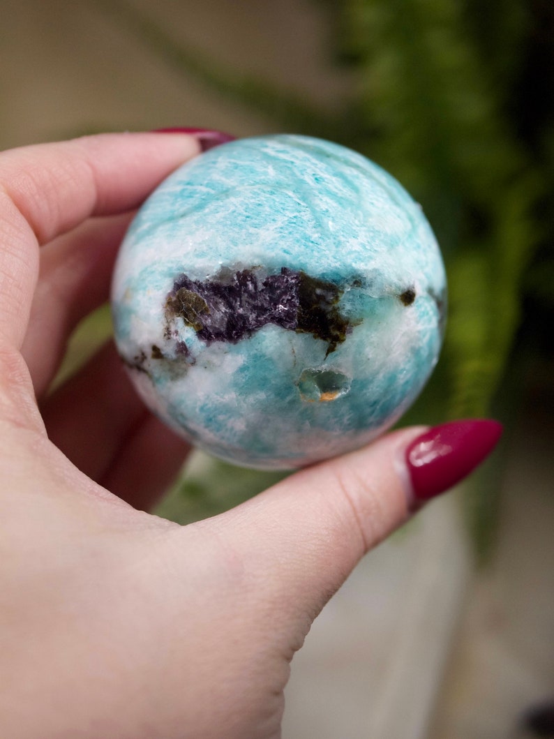 May include: A smooth, light blue and white Amazonite sphere with dark inclusions. The sphere is being held in a hand.