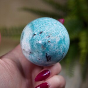 May include: A smooth, blue and white Amazonite sphere with dark inclusions. The sphere is being held in a hand with red nail polish.