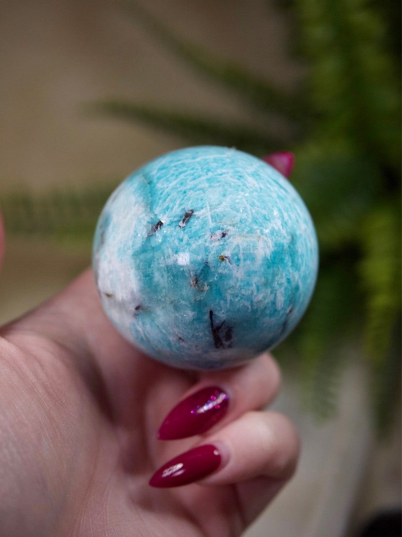 May include: A smooth, blue and white Amazonite sphere with dark inclusions. The sphere is being held in a hand with red nail polish.