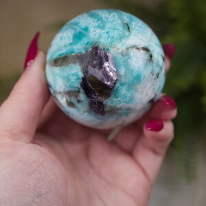 May include: A blue and green amazonite sphere with a purple amethyst inclusion. The sphere is smooth and polished.